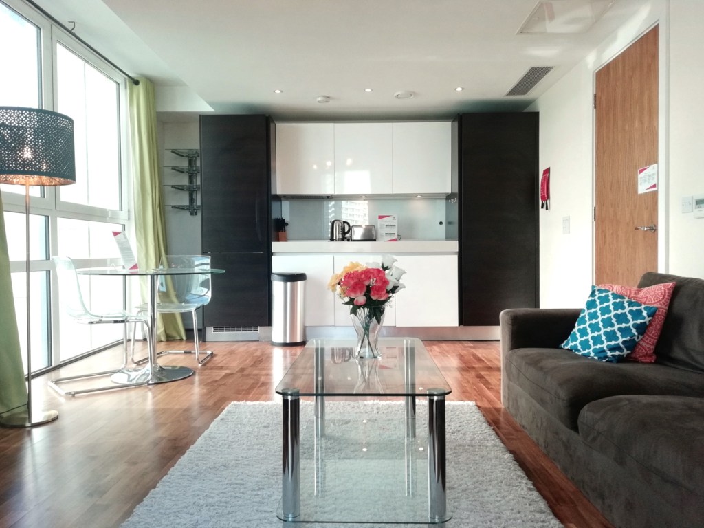 Interior view of a serviced apartment in Moorgate, showcasing a cozy living area with a dark brown sofa, glass coffee table, and a floral centerpiece. There is a kitchenette with modern appliances and a dining area with two clear chairs. Natural light enters through large windows with green curtains.