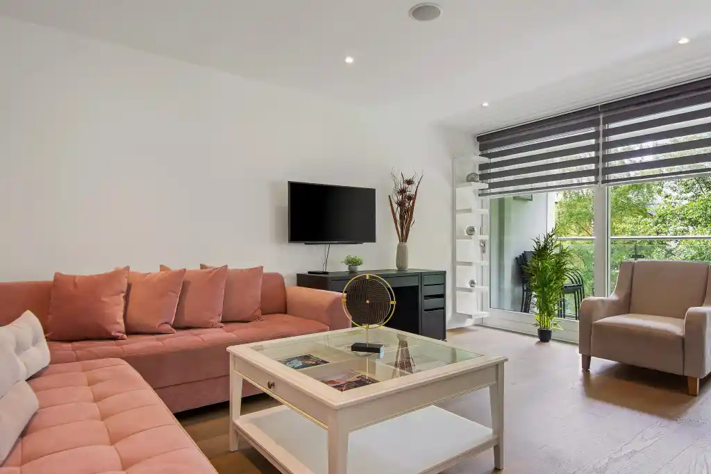 Spacious living room with pink sofa, armchair, and large window with blinds.