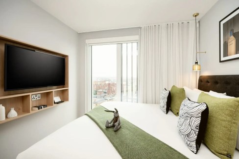 Modern hotel room with a large bed, green and black decorative pillows, and a wall-mounted flat-scre.