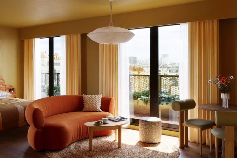 Bright hotel room with large windows and balcony overlooking Paris skyline.