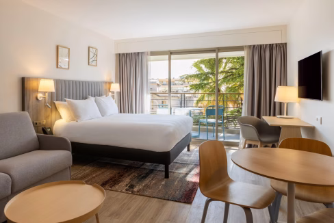 Modern hotel room in an aparthotel in Cannes featuring a large bed, a seating area with a gray couch, and a dining table with four chairs. Large windows allow natural light, with a view of palm trees outside.