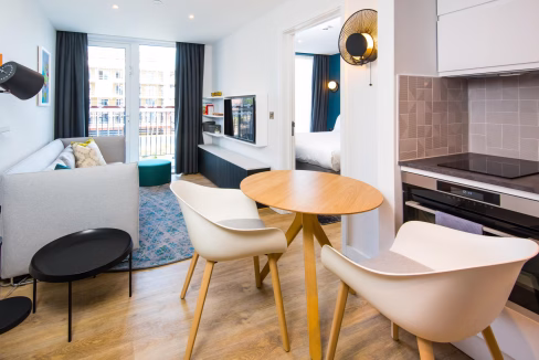 Interior view of the Cardiff Atlantic Wharf Aparthotel featuring a bright living space with a sofa, dining table, and chairs. A mounted TV and kitchenette are visible, along with large windows allowing natural light.