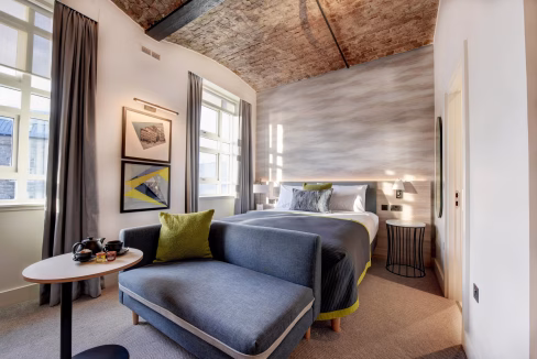 A modern room at the Dundee City Centre Aparthotel, featuring a bed with gray bedding, a cozy seating area with a green accent pillow, and large windows allowing natural light. Artwork hangs on the walls, and a small table with cups is visible.