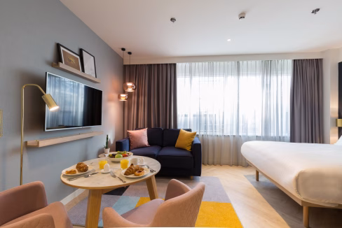 A modern hotel room at The Hague Central Aparthotel, featuring a bed, a small couch with cushions, and a round table set with breakfast items. Natural light enters through large windows with sheer curtains, enhancing the contemporary decor.