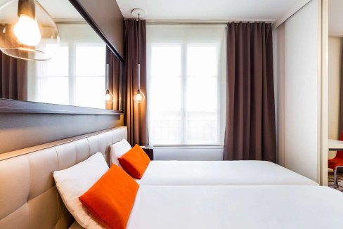 Modern hotel room with two beds, white linens, orange pillows, and contemporary lighting.