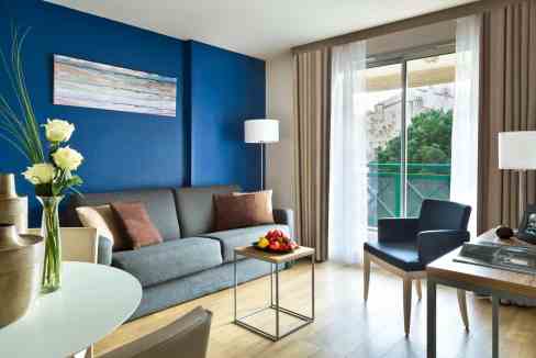 Bright hotel suite with stylish furniture, large window, and balcony overlooking greenery.