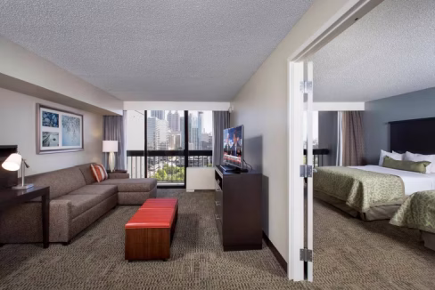 A spacious room in an aparthotel in Atlanta featuring a seating area with a sofa, an orange ottoman, a desk, and a television. Beyond an internal partition, a bedroom area with a bed and nightstands is visible, alongside city views through large windows.