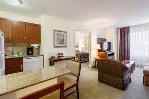An interior view of a suite in the Austin Airport Hotel, featuring a kitchenette with wooden cabinets, a dining table, and a living area with two chairs and a sofa. A bed is visible through an opening, with a TV in the living area and large windows providing natural light.