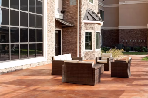 A patio area at the Austin Aparthotel featuring four wicker chairs and cushions arranged around a small table. The backdrop includes a large window with stone accents on the building, set on a wooden deck under bright sunlight.