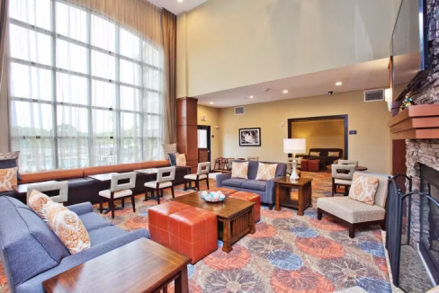 A spacious lobby area in the South Austin Aparthotel featuring large windows, a mix of seating including sofas and chairs, and a coffee table. The decor includes vibrant carpets and a stylish fireplace, creating a welcoming atmosphere.