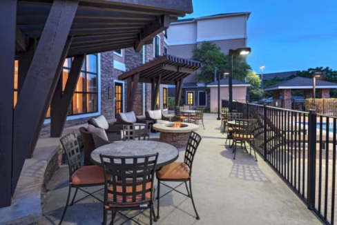 Outdoor seating area of the Northwest Austin Aparthotel, featuring round tables with chairs and a fire pit, alongside a pool. The space is illuminated by lamps and covered by wooden awnings, with trees visible in the background, suggesting a welcoming atmosphere.