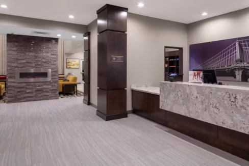 The reception area of a Cedar Park aparthotel near Austin, featuring a modern front desk made of stone, a textured stone wall with an electric fireplace, and a seating area with yellow chairs in the background.