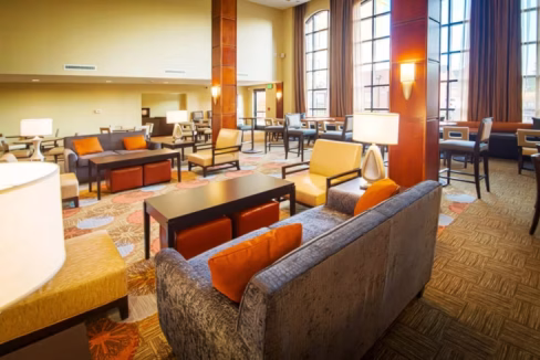 A spacious lounge area at the Denver Central Park Aparthotel featuring modern furniture. The design includes a mix of sofas and chairs, tables with lamps, and large windows providing natural light in a welcoming atmosphere.
