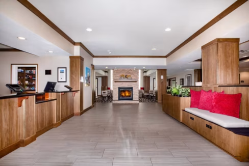 The lobby of the Denver Airport Aparthotel features a modern design with wooden accents, a seating area with red cushions, and a fireplace at the back. The reception desk is on the left with a computer and menus, creating a welcoming atmosphere.
