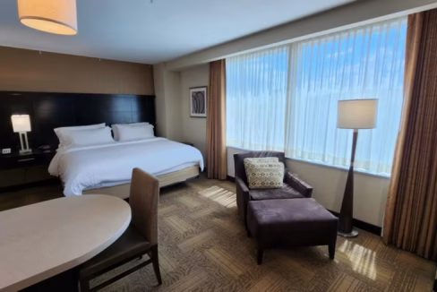 A well-lit hotel room at the Denver Downtown Aparthotel, featuring a king-size bed with white linens, a chair with a patterned cushion, a small table, and large windows letting in natural light.