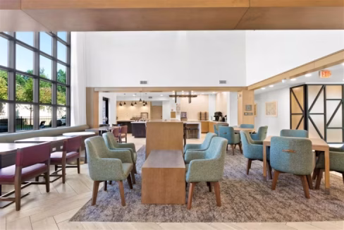 Interior view of the Atlanta Duluth Aparthotel lobby featuring modern furniture. A seating area with teal chairs and wooden tables is visible, alongside a dining area with purple chairs, and large windows allowing natural light.