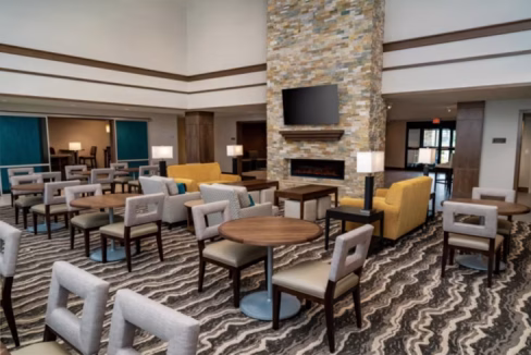 Interior of the Nashville Franklin Aparthotel featuring a spacious lounge area with beige and yellow seating, round wooden tables, and a stone fireplace. Large windows allow natural light, creating an inviting atmosphere.