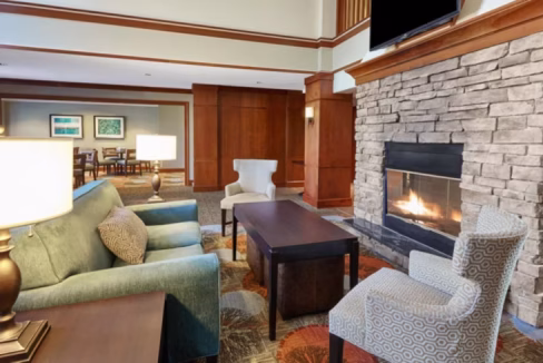 A cozy hotel lobby featuring a green sofa, a stone fireplace, and two armchairs. A coffee table is positioned in front of the sofa, and a lamp sits beside it. Warm lighting creates an inviting atmosphere, suitable for relaxing during a business trip to Chicago.