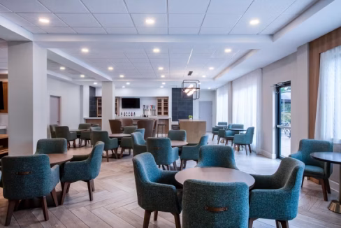 A spacious lounge area in the Atlanta Airport Hotel, featuring several round tables surrounded by teal upholstered chairs. Large windows allow natural light, and a countertop bar area is visible in the background.