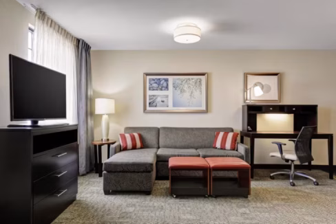 A well-furnished living area in the Washington D.C. Greenbelt Aparthotel, featuring a gray sofa with striped cushions, a TV on a black cabinet, a small table lamp, and a workspace with a desk and chair against a light-colored wall.