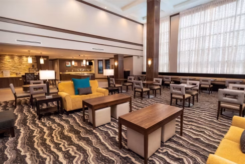 A spacious lobby area of the Washington D.C. East Aparthotel featuring modern seating arrangements with gray armchairs and yellow sofas, wooden coffee tables, and large windows allowing natural light.