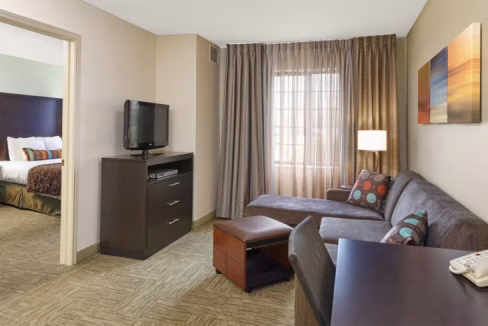 A modern living area in an aparthotel in Lincolnshire Chicago, featuring a gray couch, a coffee table, a TV on a stand, and a lamp next to a window with sheer curtains. Neutral walls and patterned carpet complete the welcoming atmosphere.
