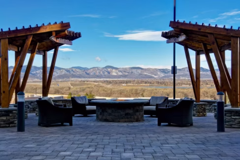 View from the Denver Highlands Ranch Aparthotel showing a stone patio with four black lounge chairs surrounding a circular fire pit, framed by two wooden pergolas. In the background, mountains extend under a clear blue sky.