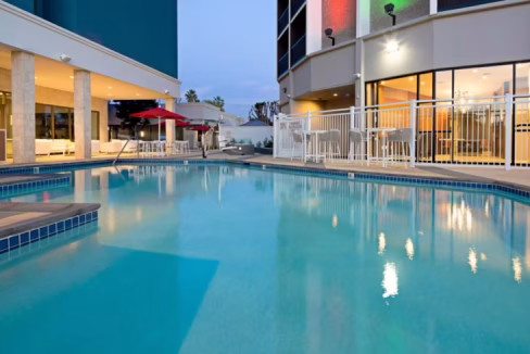 A view of a swimming pool at dusk in Long Beach Airport Aparthotel. The pool features clear water, surrounded by lounge chairs and a red umbrella. The hotel building is visible in the background, with large windows reflecting the evening light.