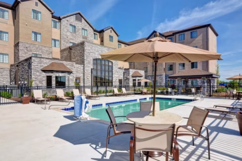 Outdoor pool area of the Mount Juliet Nashville Aparthotel, featuring sun loungers and patio umbrellas. The building has stone and stucco walls, with large windows allowing natural light. A clear blue sky completes the sunny atmosphere.