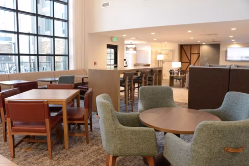 Interior view of a modern lounge area in the Nashville Murfreesboro Aparthotel. The space includes wooden tables and upholstered chairs, with large windows providing natural light. A cozy atmosphere is created by the neutral color palette and contemporary furnishings.
