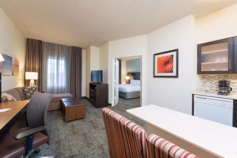 A contemporary suite in the Chicago Oakbrook Terrace Aparthotel, featuring a sitting area with a desk, a kitchenette, and a separate bedroom. Natural light filters through large windows, illuminating the modern decor and furnishings.