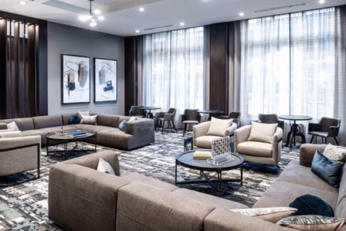 A modern lounge area at the Boston Quincy Aparthotel featuring two large, gray sectional sofas, round coffee tables, and stylish chairs. The room has large windows with sheer curtains and framed artwork on the walls, combining comfort and elegance.