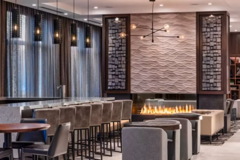 Interior of the Boston Logan Airport Aparthotel lobby featuring modern seating arrangements, a stylish bar area with high stools, and a decorative fireplace with a textured wall in the background. Ambient lighting creates a welcoming atmosphere.