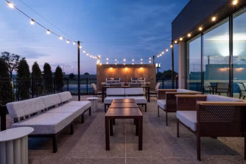 A rooftop patio of a Chicago airport hotel featuring several seating areas with gray couches and wooden tables, illuminated by string lights under a twilight sky.