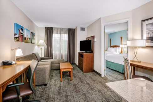An ensuite room in the Austin Round Rock Aparthotel featuring a sitting area with a gray sofa and wooden coffee table, a desk with a chair, and a flat-screen TV. The room has large windows covered by curtains, with an adjoining bedroom visible.