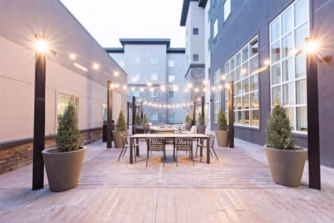A modern outdoor patio area featuring a dining table surrounded by chairs, illuminated by string lights. Potted evergreen plants are placed at either end, while the surrounding buildings are constructed with a mix of modern materials. This is part of Denver North Aparthotel.