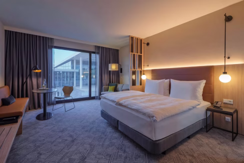 A modern hotel room with a large bed featuring white linens, a wooden desk, and a small table with a chair. Floor-to-ceiling windows showcase outside views, and soft lighting enhances the space. The room is part of the Cologne Messe Auenweg Aparthotel.