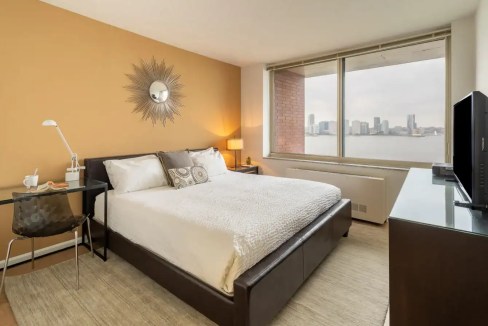 Cozy bedroom with large window overlooking Battery City Park skyline.