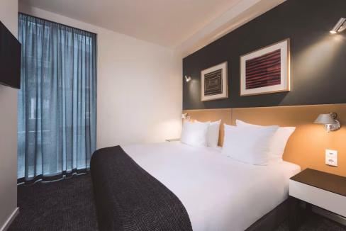 A modern hotel room featuring a large bed with white pillows and a dark bedspread. The walls are adorned with framed artwork, and a window dressed with sheer blue curtains allows natural light to enter, highlighting the contemporary decor of the Copenhagen Aparthotel.