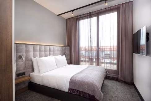 A contemporary hotel room featuring a queen-size bed with white linens and a gray accent blanket. Large windows with sheer curtains offer natural light, and a wall-mounted TV is visible across from the bed. This room is part of the Nuremberg Lorenz Aparthotel.