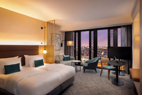 A modern hotel room in the Munich Au-Haidhausen Aparthotel featuring two beds with white linens and teal accent pillows. Large windows provide a view of the city, and the room includes a seating area with a couch, table, and a television.