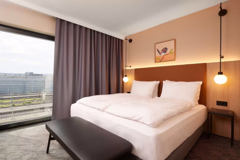 A modern hotel room at the Dusseldorf Harkortstraße Aparthotel featuring a large bed with white bedding, a dark bench, and minimalistic furnishings. Floor-to-ceiling windows reveal an urban landscape with soft curtains and stylish lighting fixtures above the bed.