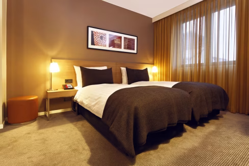 A hotel room featuring two twin beds with dark brown bedding arranged neatly. A bedside table with a lamp and a phone is beside the beds. Warm curtains frame a large window, while a small round orange ottoman is placed in the corner. The setting is part of the Robert Koch Platz Aparthotel.