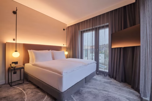 A modern hotel room featuring a large, white bed with two cozy pillows, situated beside a small nightstand with a lamp. Soft curtains drape over a large window, and a flat-screen TV is mounted on the wall. This is a room in the Wiesbaden Taunusstrasse Aparthotel.