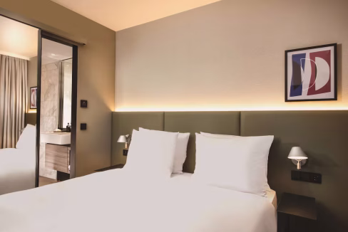 A modern hotel room featuring a double bed with white pillows and a dark green headboard. On the wall, there are two framed abstract prints and a mirror reflecting the light fixtures. The bathroom is visible through the glass door.