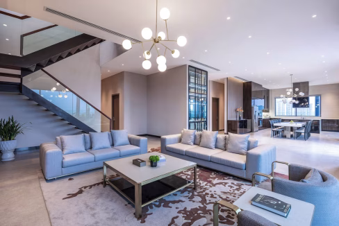 A modern living area in the Hanoi Xuan Dinh Aparthotel featuring light gray sofas arranged around a coffee table. A decorative plant sits beside a glass staircase, and the space is illuminated by stylish light fixtures. The kitchen area is visible in the background.