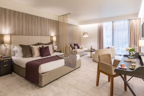 An elegantly furnished hotel room at Swiss Cottage Aparthotel features a king-size bed with purple and white bedding, a seating area with a light gray sofa, and a wooden desk with a flower vase. Large windows allow natural light into the room.