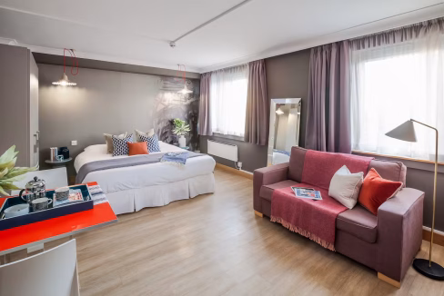 A modern guest room at the Watford Junction Aparthotel featuring a double bed with decorative pillows, a pink sofa, and large windows with sheer curtains. A small table with refreshments is visible beside a mirror.