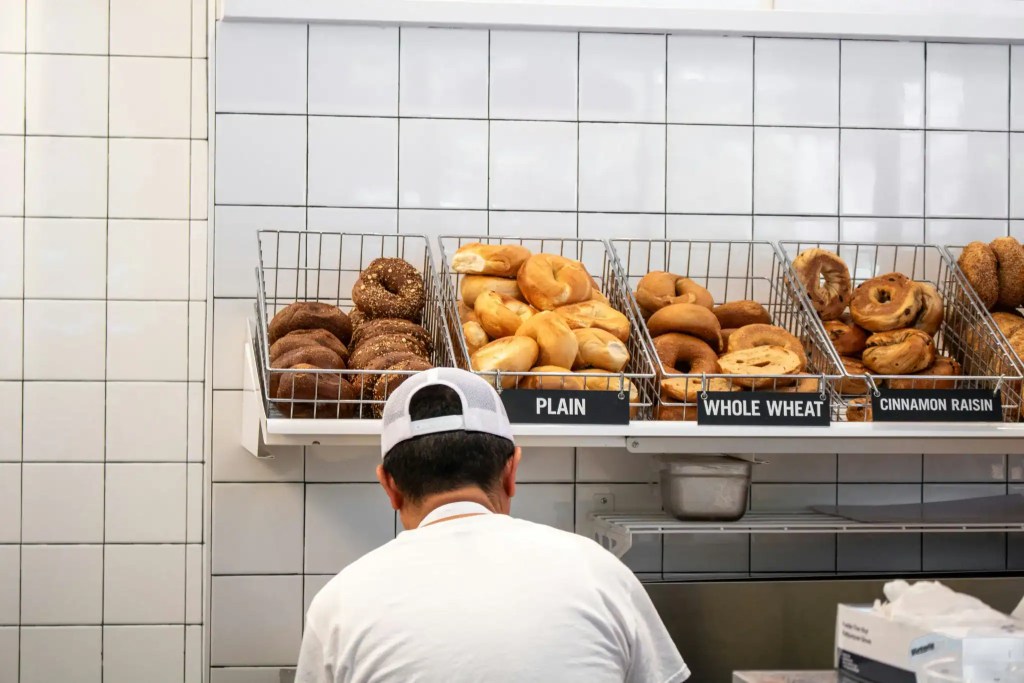 Assorted bagels on display in a bakery with labels for plain, whole wheat, and cinnamon raisin.