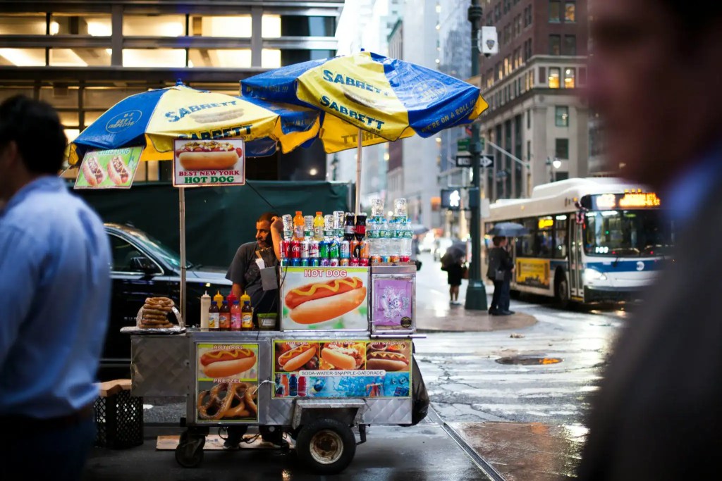 Hot dog cart with umbrellas in New York City, bustling urban scene.